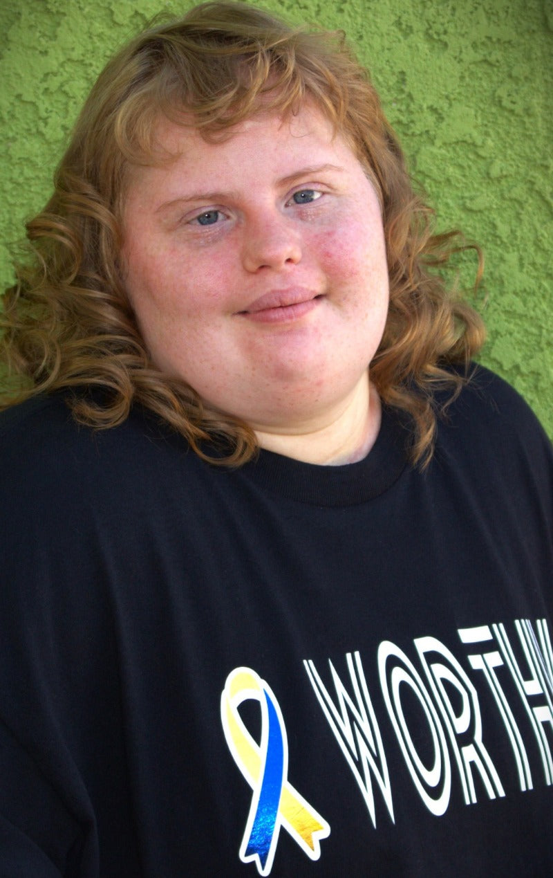 A young woman, soft round face, and soft beautiful hair wearing a black tee-shirt. The word, "Worthy is emblazoned across the front next to a Down Syndrome Awareness ribbon of blue and yellow.